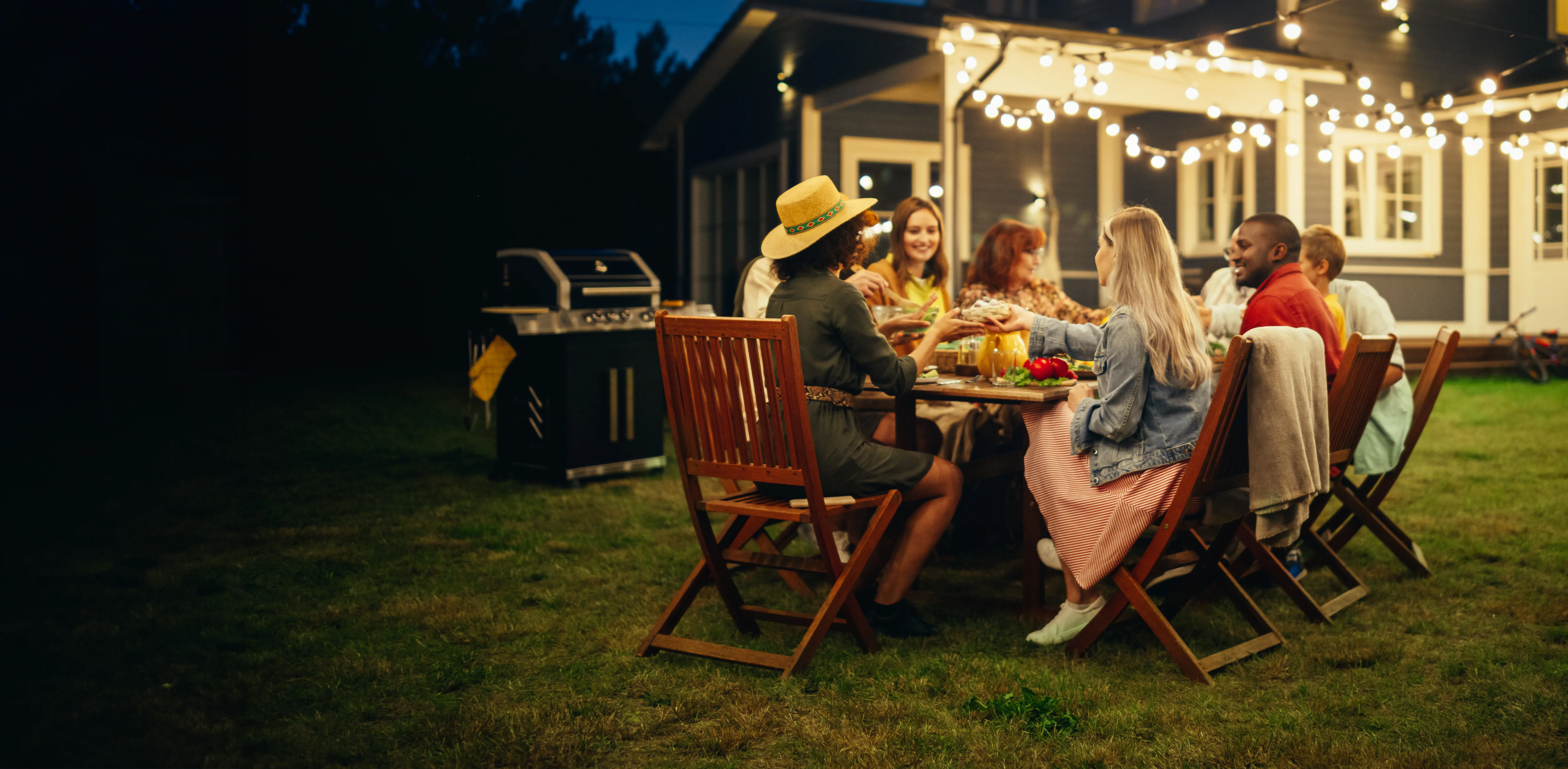 A group of people sit around a table outdoors at night, sharing a meal under string lights in a backyard near a house. The scene is warm and festive, with food and drinks on the table.