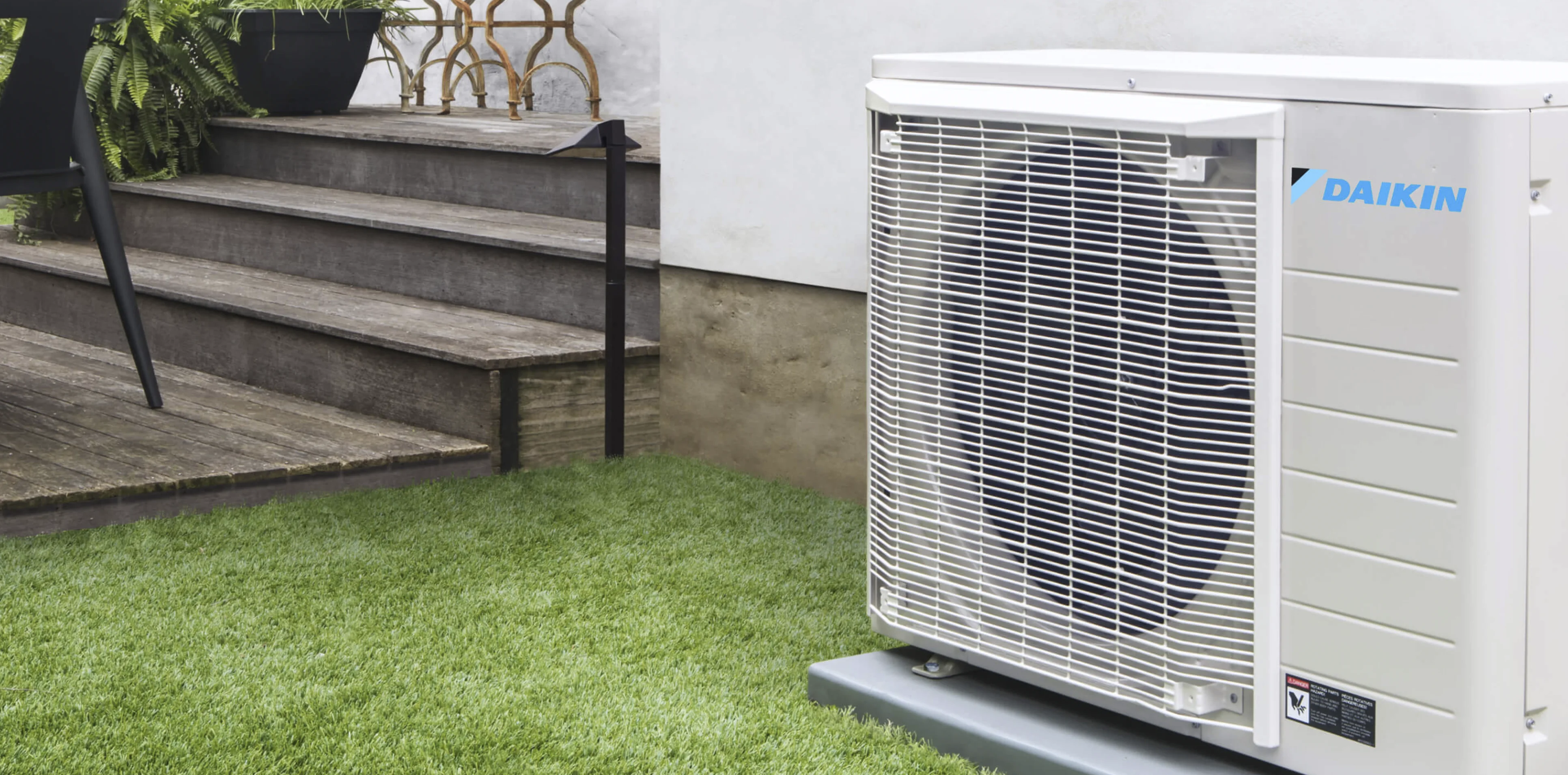 An outdoor Daikin air conditioning unit is placed on a concrete slab next to a modern wooden staircase and green artificial grass in a garden or backyard setting.