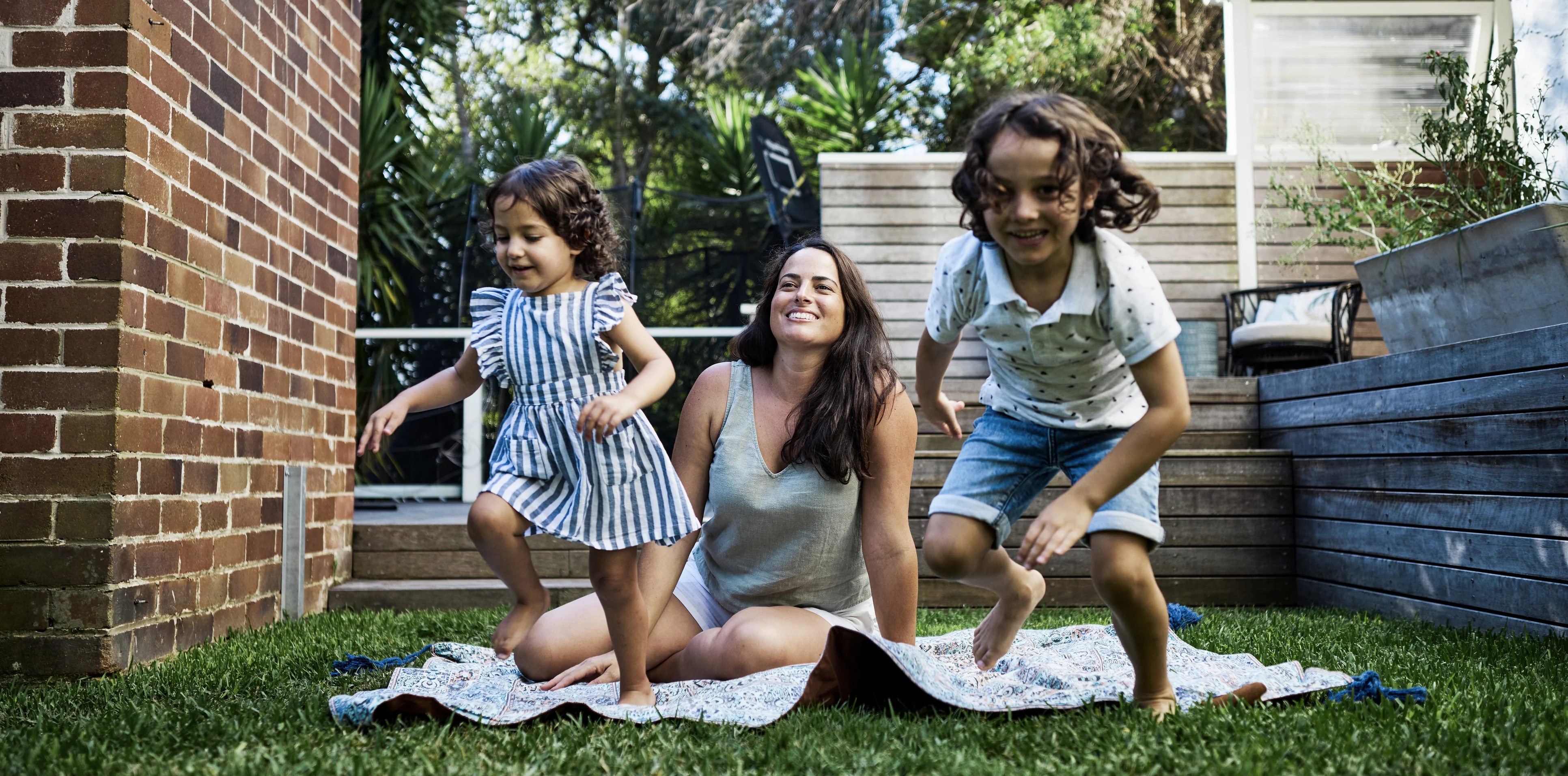 A woman sits on a blanket in the grass, smiling as two children joyfully jump around her in a backyard with trees, a wooden deck, and a brick wall.
