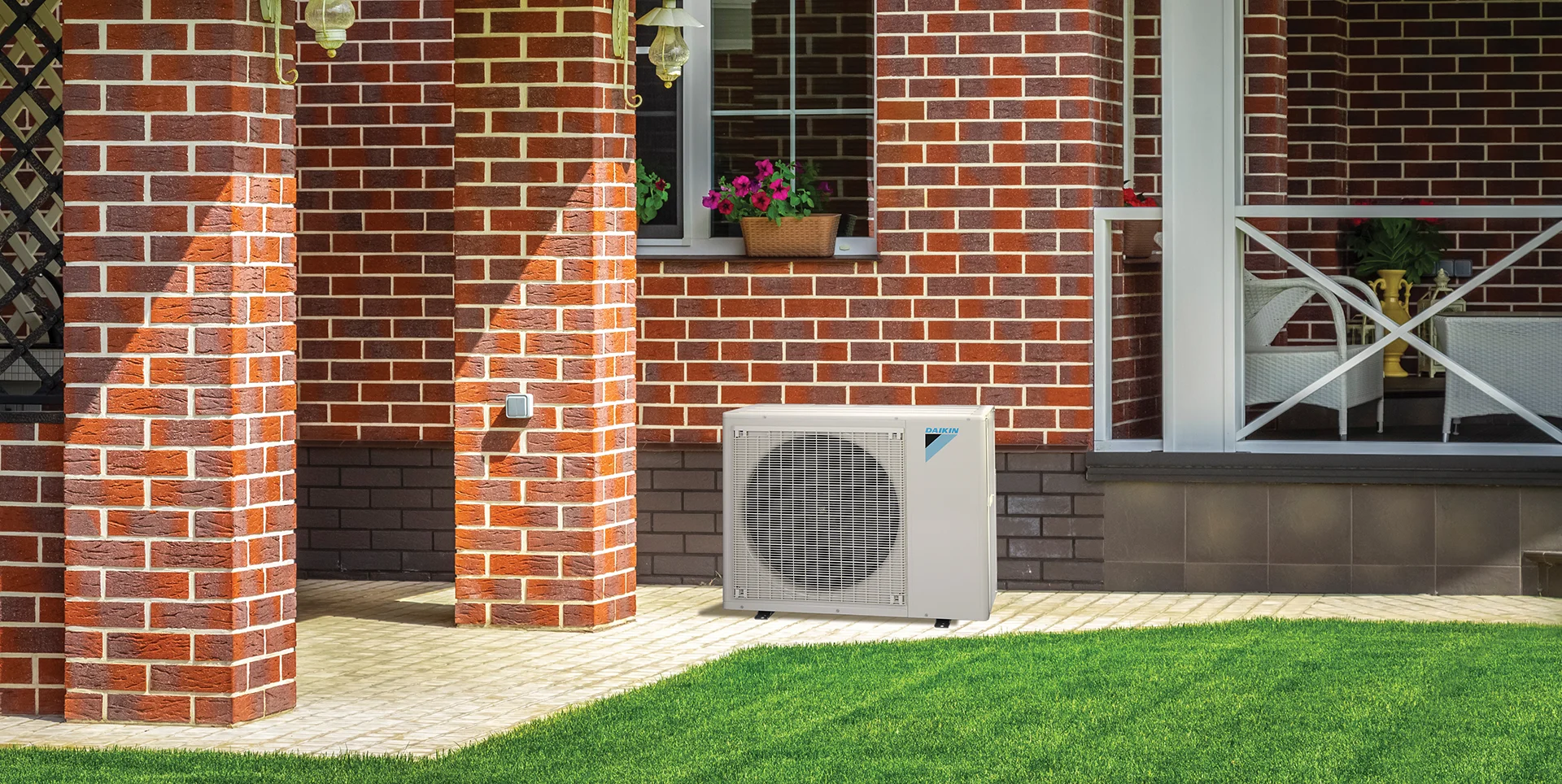 A heat pump unit sits on a paved area next to a red brick house with white trim, green grass, and potted flowers on a windowsill.