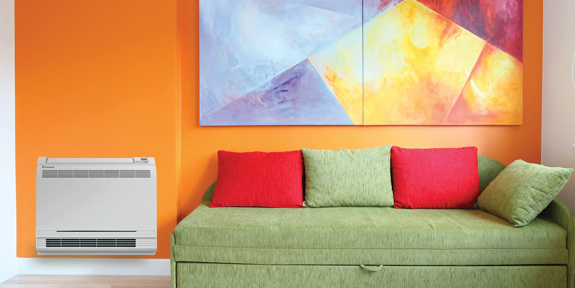 A bright living room with an orange wall, a green sofa with red cushions, a colorful abstract painting above the sofa, and a white wall-mounted air conditioner to the left.