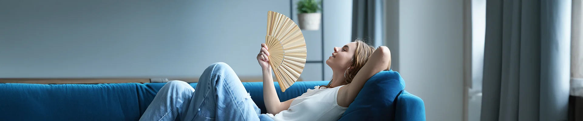 A woman relaxes on a blue couch, leaning back with her eyes closed while fanning herself with a handheld fan in a bright, modern living room.