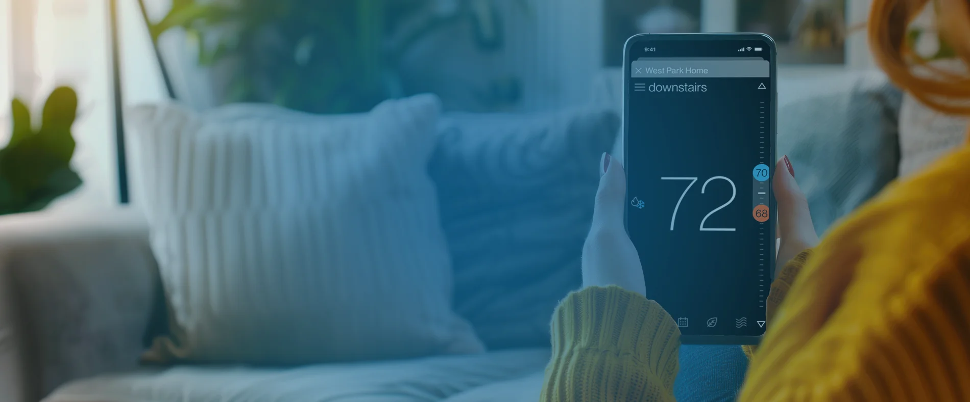 A person in a yellow sweater holds a smartphone displaying a smart thermostat app set to 72 degrees, sitting in a cozy living room with a sofa and green plants in the background.